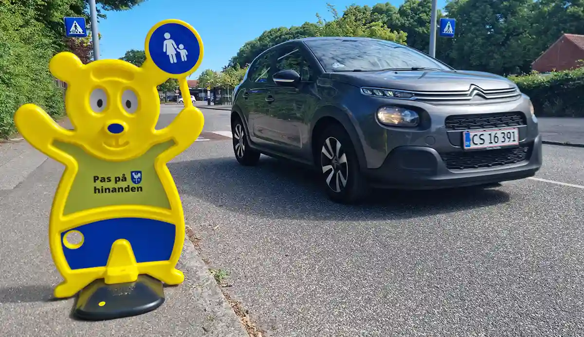 Sikker skolevej. En bil kommer kørende. Ved siden af vejen er en Trafik Bjørn. Det er et nudge. Den kaldes også for Trafic Teddy. Den sælges af firmaet Brave.