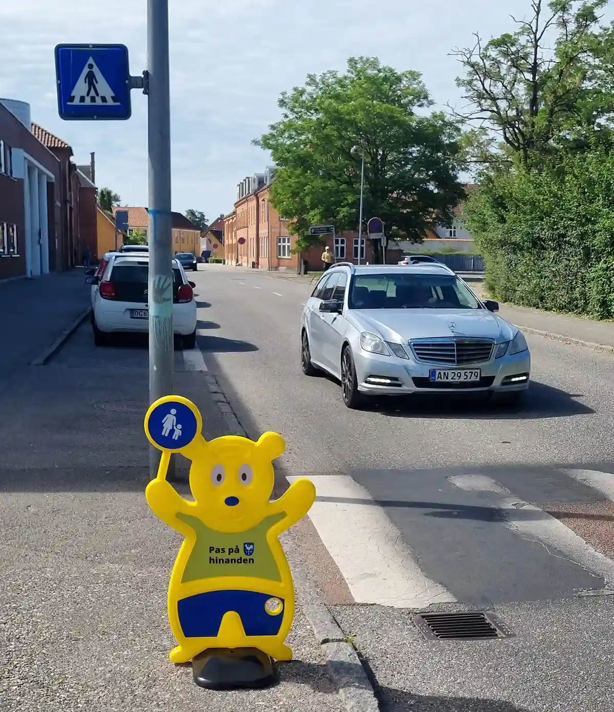 Sikker skolevej. Ved fodgængerfelt. Trafik Bjørn skaber opmærksomhed. Der er en Mercedes bil lige ved fodgængerfeltet ved skolen. Ved siden af står en gul Trafik Bjørn fra Brave. Det er et nudge. Trafik Bjørn skaber opmærksomhed.