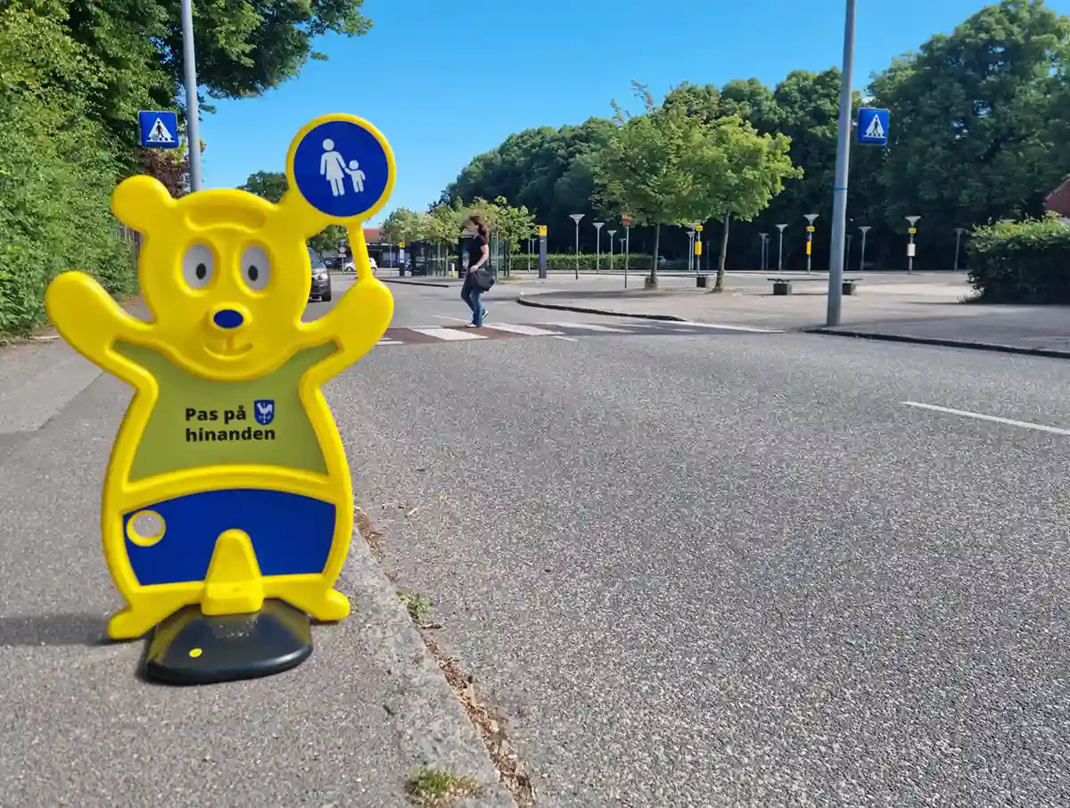 Sikker skolevej. Ved fodgængerfelt. Skoleelever går over fodgængerfelt. Ved siden af står en gul Trafik Bjørn fra Brave. Det er et nudge. Trafik Bjørn skaber opmærksomhed.
