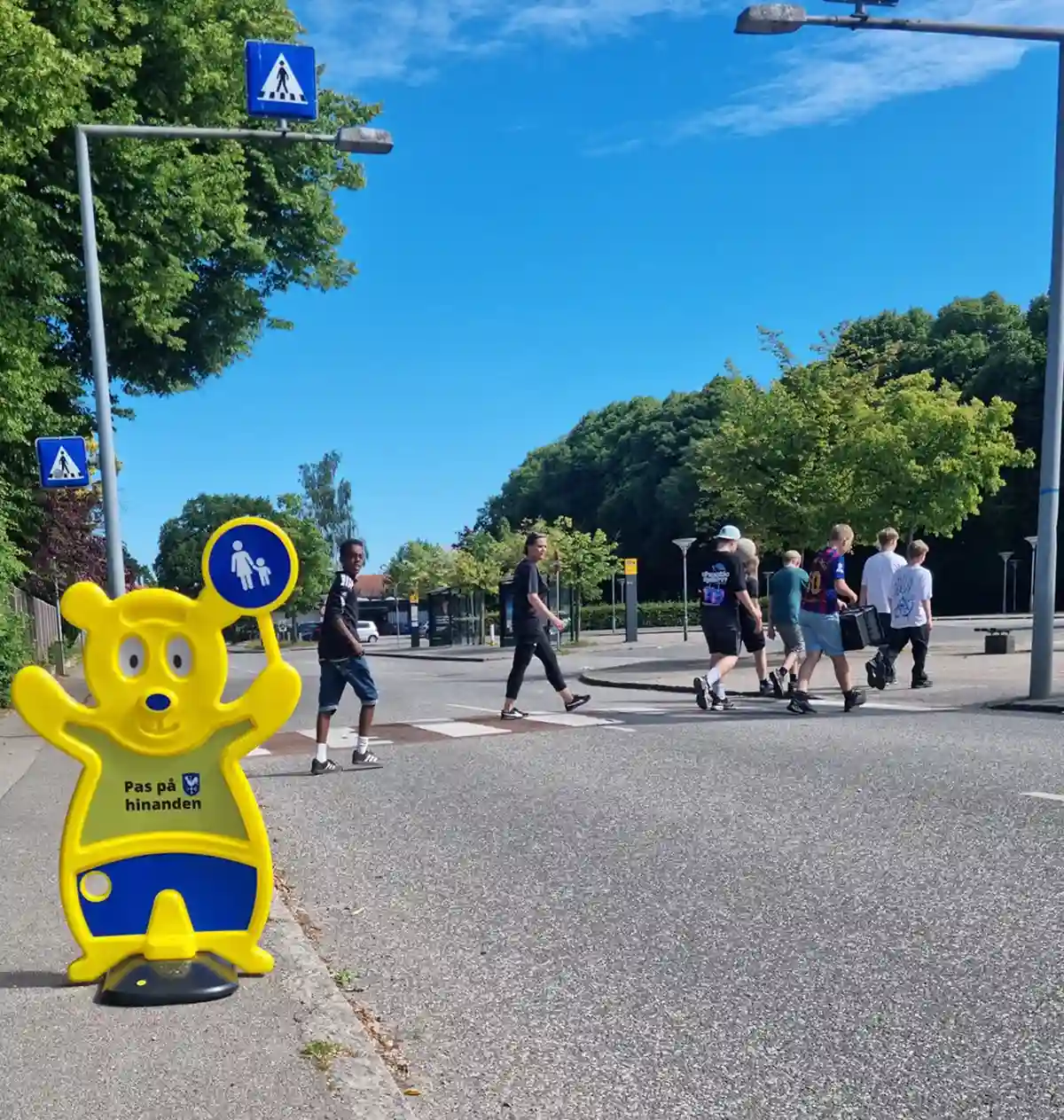 Nudging af elever. Ved fodgængerfelt. Skoleelever går over fodgængerfelt. Ved siden af står en gul Trafik Bjørn fra Brave. Det er et nudge. Trafik Bjørn skaber opmærksomhed.